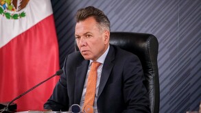 Pablo Lemus vestido de traje con corbata de naranja durante una conferencia de prensa en Jalisco y con una bandera de México detrás.