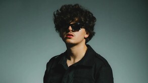 El cantante de música mexicana Gabito Ballesteros posa con cabello rizado oscuro, chaqueta negra, lentes de sol rectangulares y labios pintados de rojo.