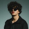 El cantante de música mexicana Gabito Ballesteros posa con cabello rizado oscuro, chaqueta negra, lentes de sol rectangulares y labios pintados de rojo.