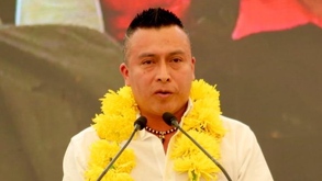 Hombre hablando al micrófono con plumas amarillas puestas en su cuello.