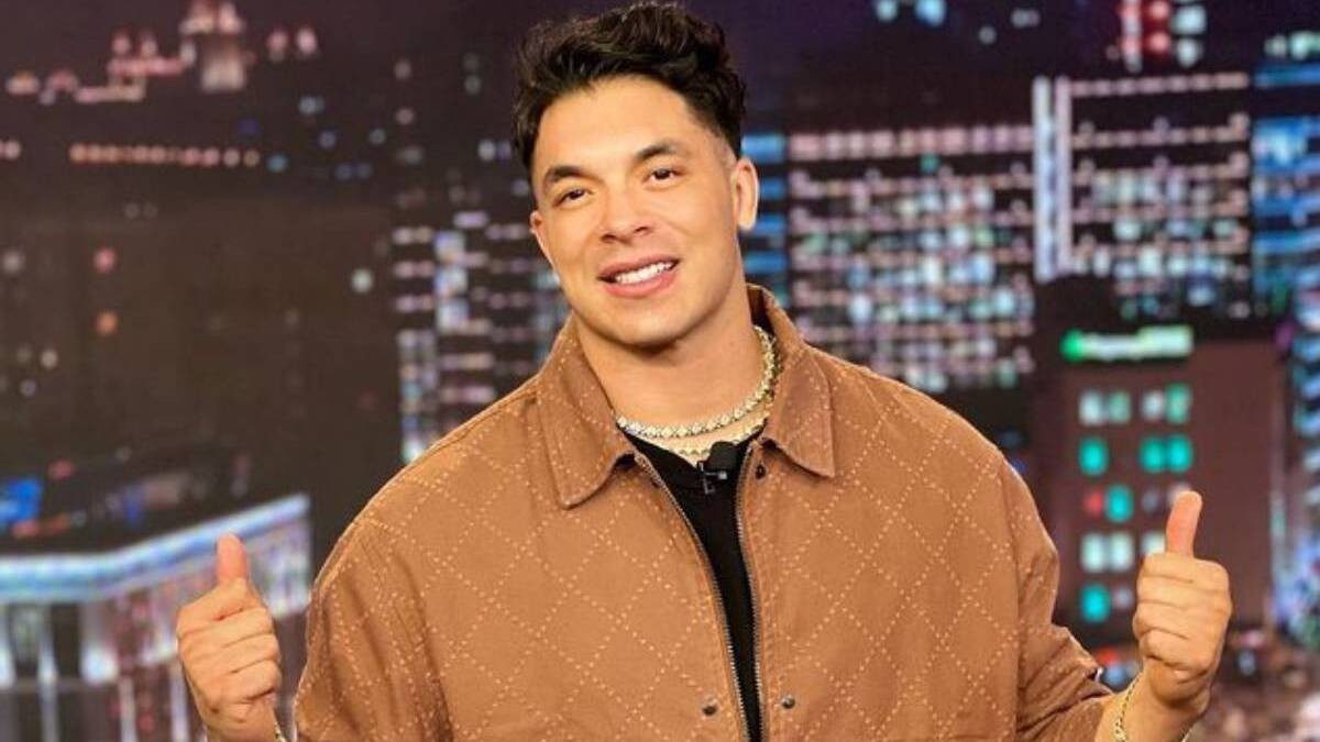 Jawy Méndez sonriendo con los pulgares levantados portando una chamarra en color cafe en un fondo que podría ser una ciudad.