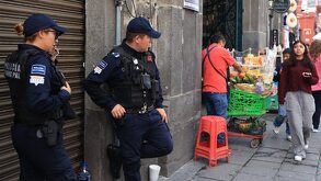 Dos hombres vestidos con uniforme de policías, recargados sobre una pared y observando a un vendedor ambulante en calles de Puebla.