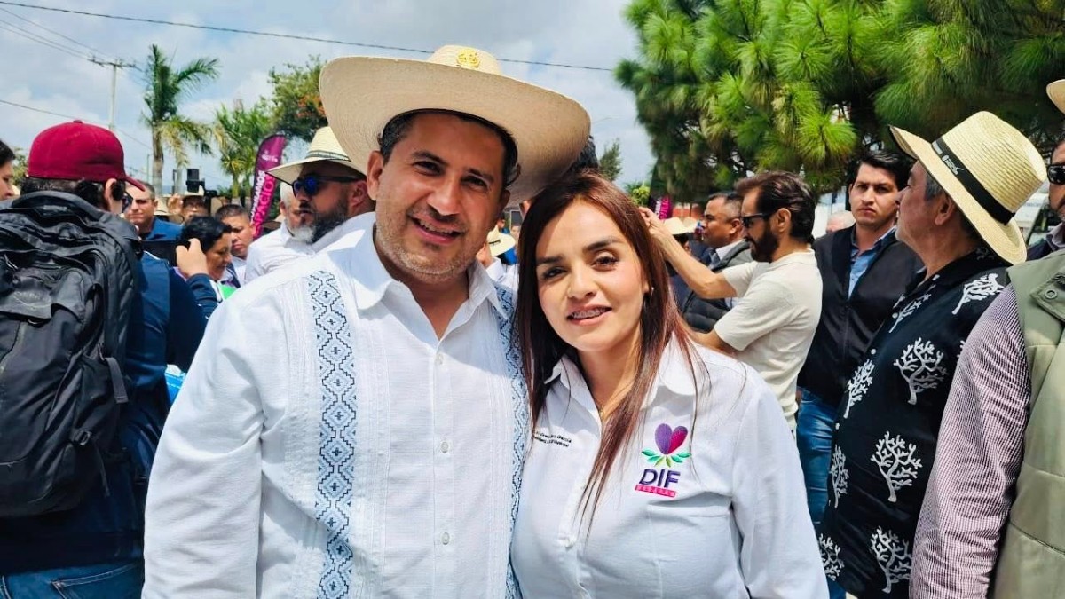 Grecia Quiroz abrazando a su esposo Carlos Manzo ambos vestidos con camisas blancas y rodeados de personas durante presentación pública en Uruapan