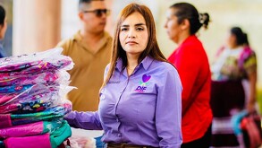 Grecia Quiroz con camisa morada del DIF junto a una pila de mochilas y con personas de fondo