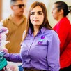 Grecia Quiroz con camisa morada del DIF junto a una pila de mochilas y con personas de fondo