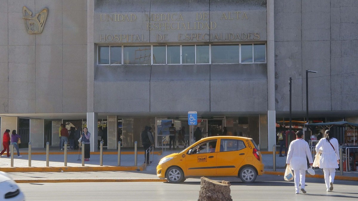 Partes frontal de clínica 71 del IMSS en Torreón. Un taxi amarillo a las afueras. Enfermeros cruzando la calle. Gente caminando rumbo a la puerta del edificio.