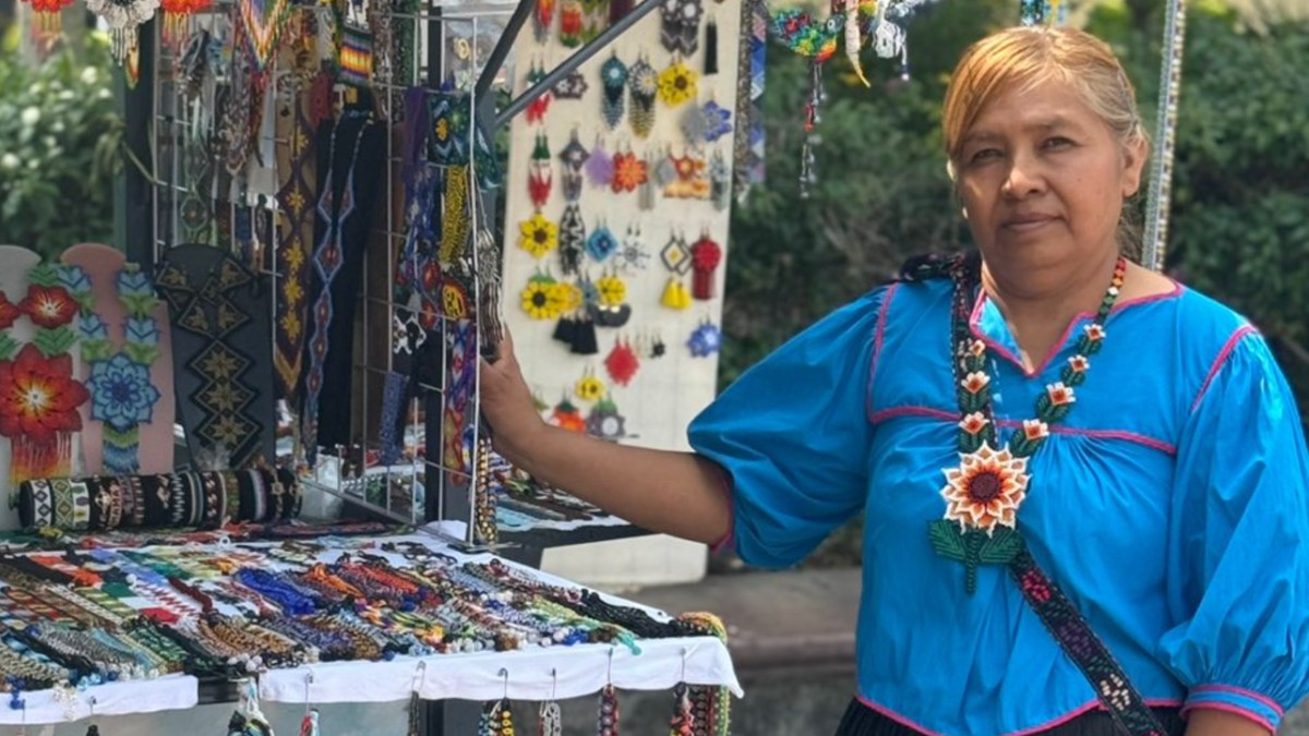 Mujer wixárika Alma Edith López al lado de su módulo de venta de distintas artesanías elaboradas con chaquira de colores en Zapopan