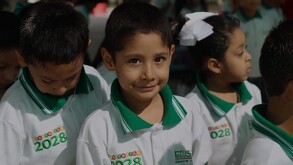 Niño sonríe mirando a la cámara con uniforme de escuela SER. A sus lados se encuentran otros niños portando el mismo uniforme y no miran la cámara.
