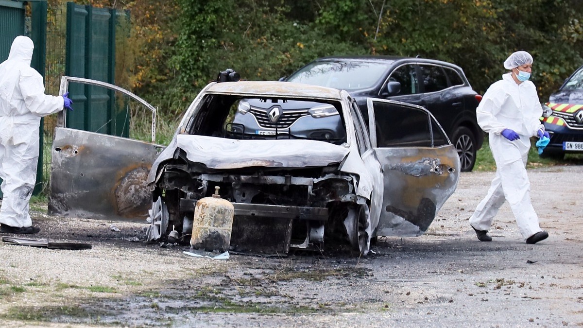 Carro incendiado con las puertas abiertas y dos personas cubiertas de ropa blanca