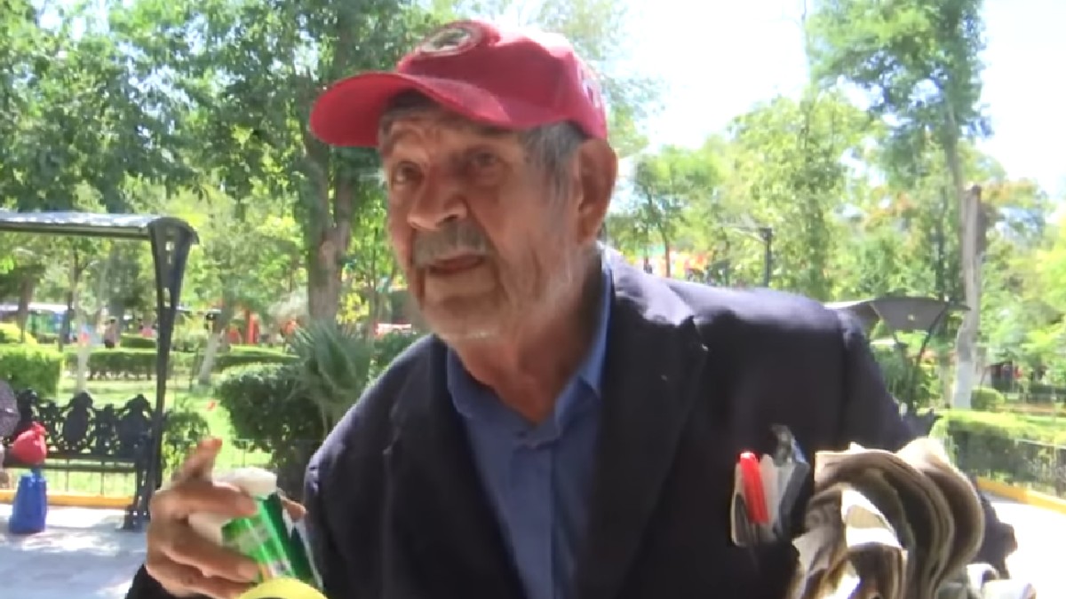 Hombre de la tercera edad con chamarra azul y gorra roja cargando una lata de refresco