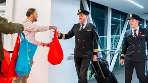 Pilotos de avión saludan y reciben bolsas de regalo rojas y azules de un promotor en la pasarela de un aeropuerto. Esto probablemente marca la inauguración de n