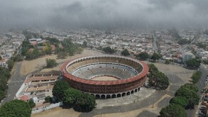Vista aérea de la Plaza de Toros El Nuevo Progreso de Guadalajara, rodeada de áreas de estacionamiento y casas. El cielo está nublado y una ligera lluvia cae so