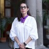 Diana Vargas, directora del Dif Jalisco usando camisa blanca y en el cuello un pañuelo morado con pilares de concreto y plantas detrás