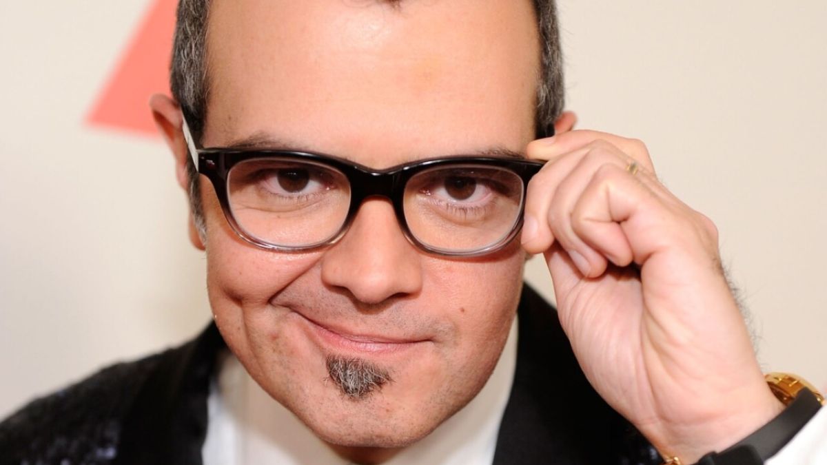 Aleks Syntek aparece sonriendo, mientras toma sus lentes con una mano, además, se le alcanza a ver un reloj en color oro con correa negra.