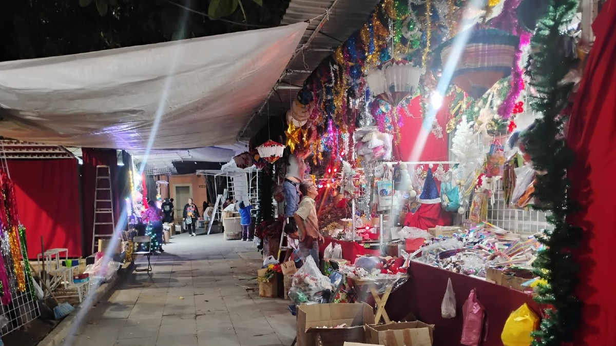 Tianguis o mercado nocturno con un pasillo cubierto por una lona blanca. Puestos llenos de adornos navideños, luces de colores y guirnaldas están a la venta con