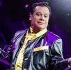 Juan Gabriel en un concierto abriendo los brazos y sosteniendo el micrófono. juan Gabriel aparece vestido de negro con dorado.
