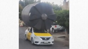 Un taxi Nissan blanco y amarillo lleva un enorme tinaco de agua negro atado en el techo como flete. El tinaco es mucho más grande que el vehículo, en una calle