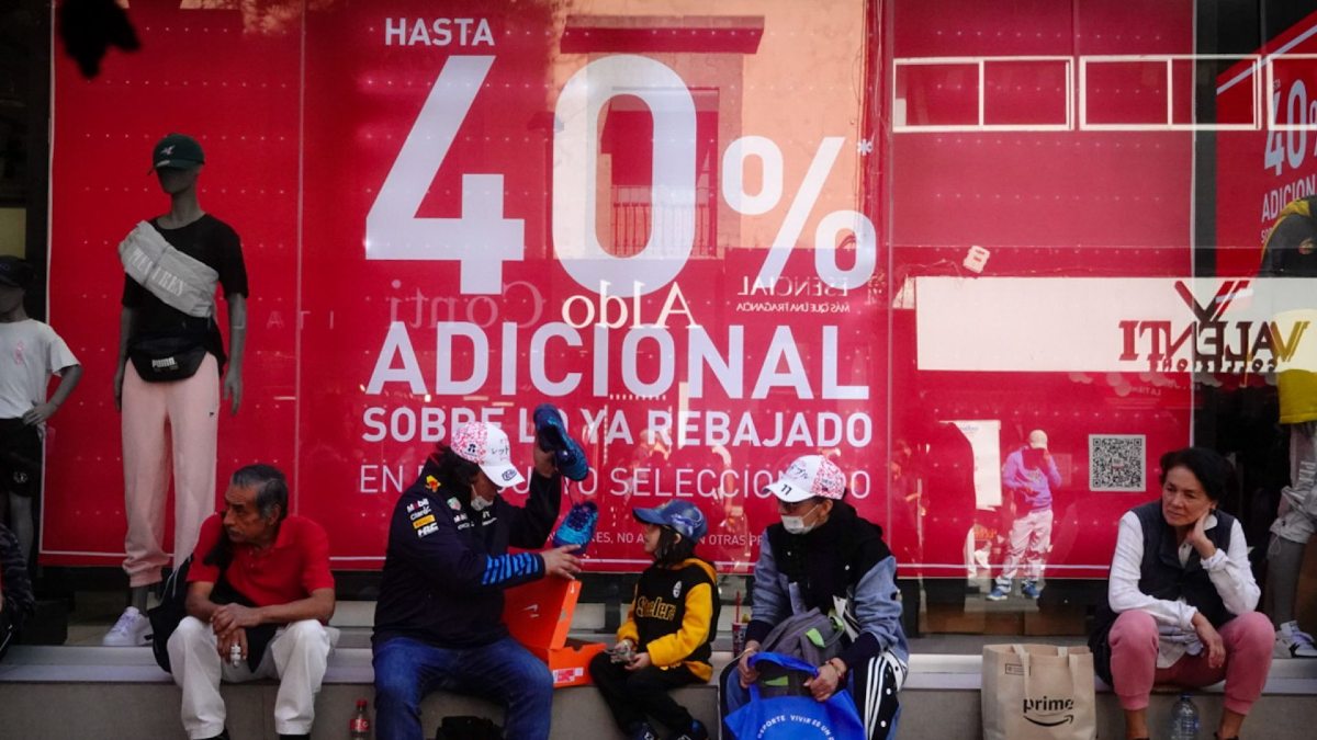 personas con bolsas de compras esperan afuera de una tienda con un anuncio del buen fin