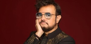 Aleks Syntek haciendo una pose colocando su mano en la barbilla, además de portar sus icónicos lentes. El cantante viste con un saco color negro.