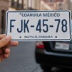 Unas placas con las letras FJK-45-78 y detrás se ve un automóvil en la ciudad de Saltillo, ya que al fondo se ve la Catedral.