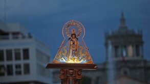La imagen de la Virgen de Zapopan (La Generala) ricamente vestida con una corona y un manto azul y dorado, es llevada en procesión por una calle con arquitectur