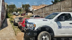 Una camioneta blanca del Instituto Jalisciense de Ciencias Forenses (IJCF) está estacionada en una calle de terracería con casas, escombros y coches viejos aban