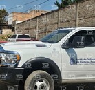Una camioneta blanca del Instituto Jalisciense de Ciencias Forenses (IJCF) está estacionada en una calle de terracería con casas, escombros y coches viejos aban