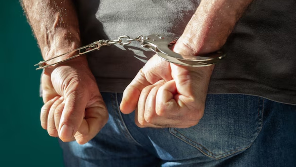 Hombre detenido con las puños cerrados en la espalda y con esposas colocadas en las muñecas usando jeans y una playera gris