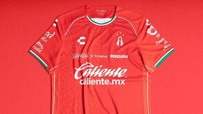 Primer plano de la nueva camiseta de fútbol de local del Atlas (roja). Tiene patrocinadores como Caliente.mx y Charly, y detalles en blanco y verde oscuro