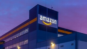 Vista exterior nocturna o al atardecer de un gran centro de distribución o almacén de Amazon. El logo de la compañía brilla sobre el edificio de color azul mari