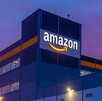 Vista exterior nocturna o al atardecer de un gran centro de distribución o almacén de Amazon. El logo de la compañía brilla sobre el edificio de color azul mari