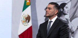 Vista de perfil de Omar García Harfuch vistiendo un traje y corbata durante conferencia de prensa con bandera de México de fondo