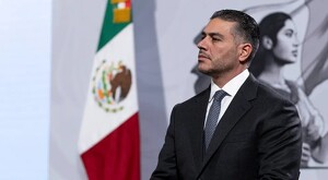 Vista de perfil de Omar García Harfuch vistiendo un traje y corbata durante conferencia de prensa con bandera de México de fondo