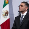 Vista de perfil de Omar García Harfuch vistiendo un traje y corbata durante conferencia de prensa con bandera de México de fondo
