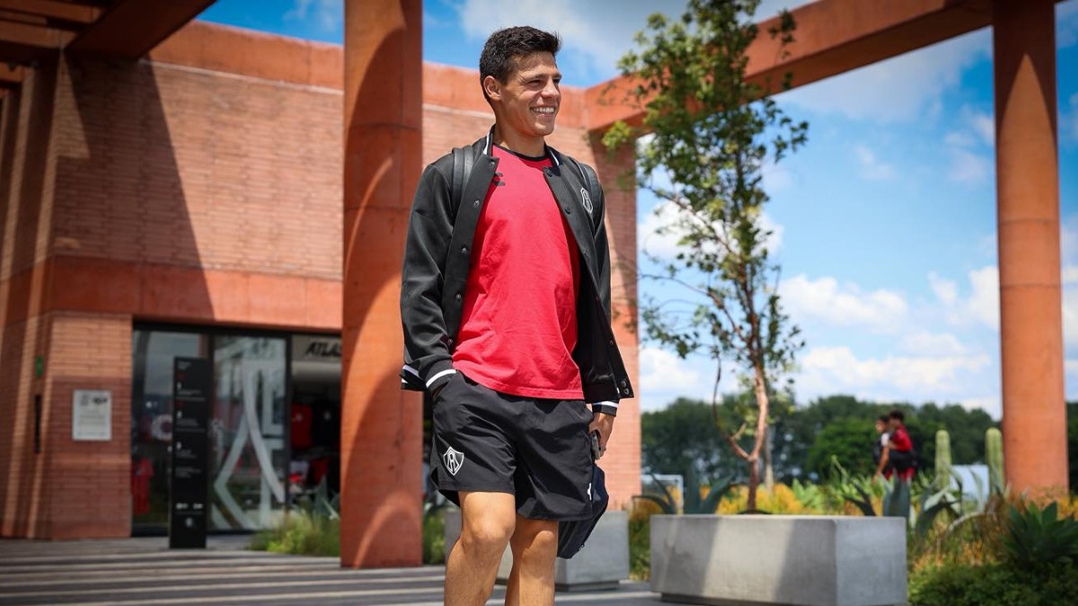 El jugador de fútbol Ponchito González camina sonriente con una camiseta roja y una chaqueta negra en un día soleado, en un campus de ladrillo