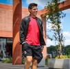 El jugador de fútbol Ponchito González camina sonriente con una camiseta roja y una chaqueta negra en un día soleado, en un campus de ladrillo
