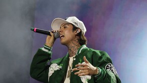 El cantante de corridos tumbados, Natanael Cano, canta en un concierto. Viste una gorra, una chaqueta verde y muestra tatuajes en el cuello. Sostiene un micrófo