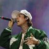 El cantante de corridos tumbados, Natanael Cano, canta en un concierto. Viste una gorra, una chaqueta verde y muestra tatuajes en el cuello. Sostiene un micrófo