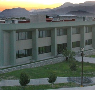 Hay un edificio color gris con unos árboles a los costados y zacate en ciertas áreas.