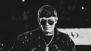 Retrato en blanco y negro del cantante de corridos tumbados, Junior H, en el escenario. Viste gafas de sol, una chaqueta con adornos y luce tatuajes en el cuell