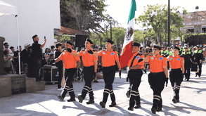 Seis elementos de protección civil o bomberos marchan en uniforme naranja y gorras negras, llevando una gran bandera de México. Están en un desfile en un día so