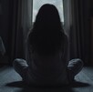 Una niña con cabello largo está sentada en el piso frente a una ventana y en un cuarto oscuro.