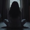 Una niña con cabello largo está sentada en el piso frente a una ventana y en un cuarto oscuro.