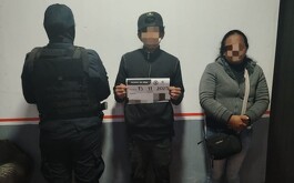 Policía de espaldas, joven y mujer parados, el joven sostiene una hoja