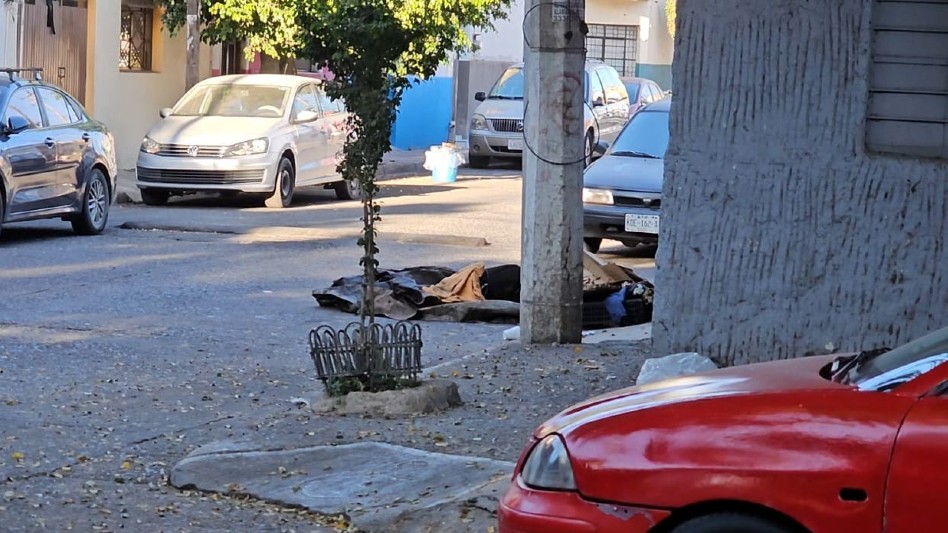 Bulto con silueta humana envuelto en cobijas y cubierto con cartones, basura, plásticos y maderas detrás de una cinta policiaca
