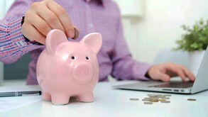 Persona metiendo monedas dentro de una alcancía en forma de cerdito rosa sobre un escritorio con una laptop y una planta a un lado