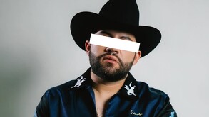 Luis R Conriquez viendo de frente y con los ojos censurados vistiendo una camisa azul y sombrero negro frente a un fondo blanco