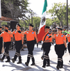 Seis elementos de protección civil o bomberos marchan en uniforme naranja y gorras negras, llevando una gran bandera de México. Están en un desfile en un día so