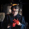 El director de cine Guillermo del Toro mira sus manos, que están cubiertas de pintura roja brillante (simulando sangre), en un ambiente oscuro y dramático, posi