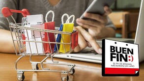 Carrito de compras con cajas y bolsas dentro sobre una mesa de madera con una laptop y una persona sosteniendo un teléfono de fondo